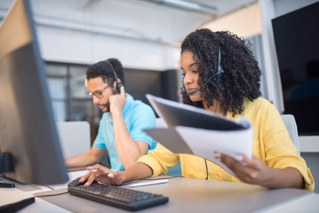 Two agents in headsets reviewing a printed document at a workstation