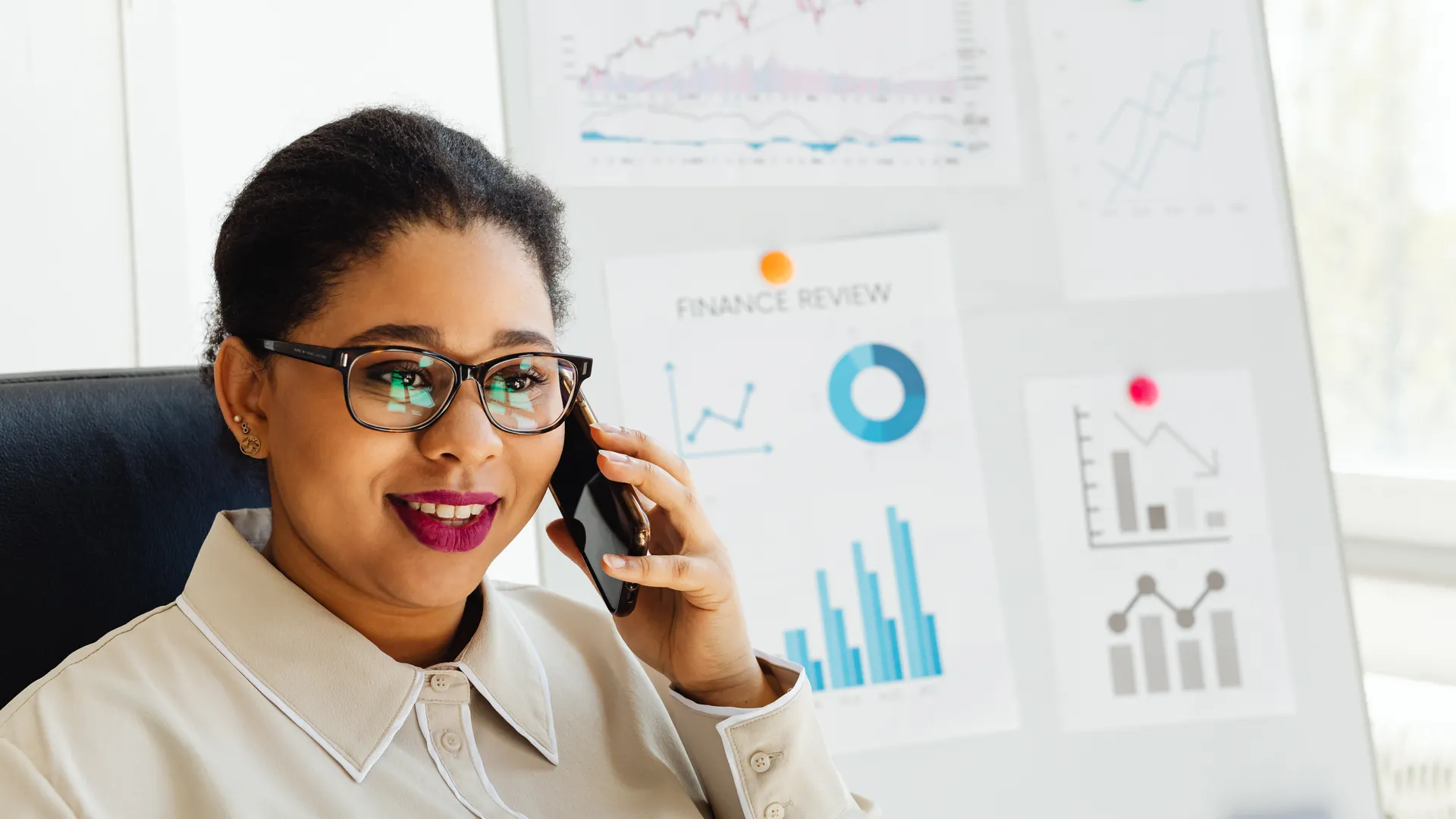 Confident businesswoman wearing glasses studies financial charts while talking on smartphone