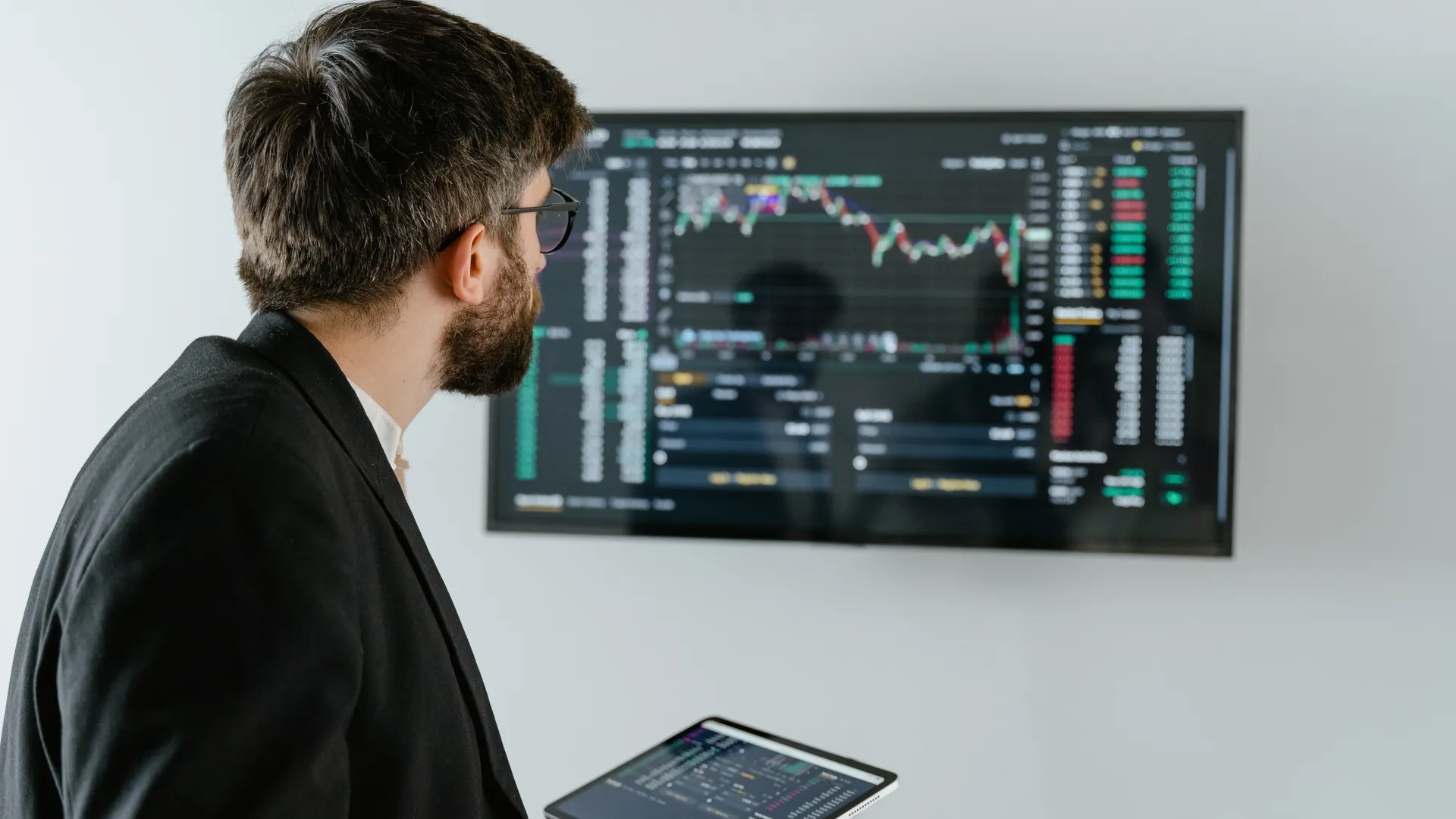A businessman examines stock market data displayed on a monitor, holding a tablet