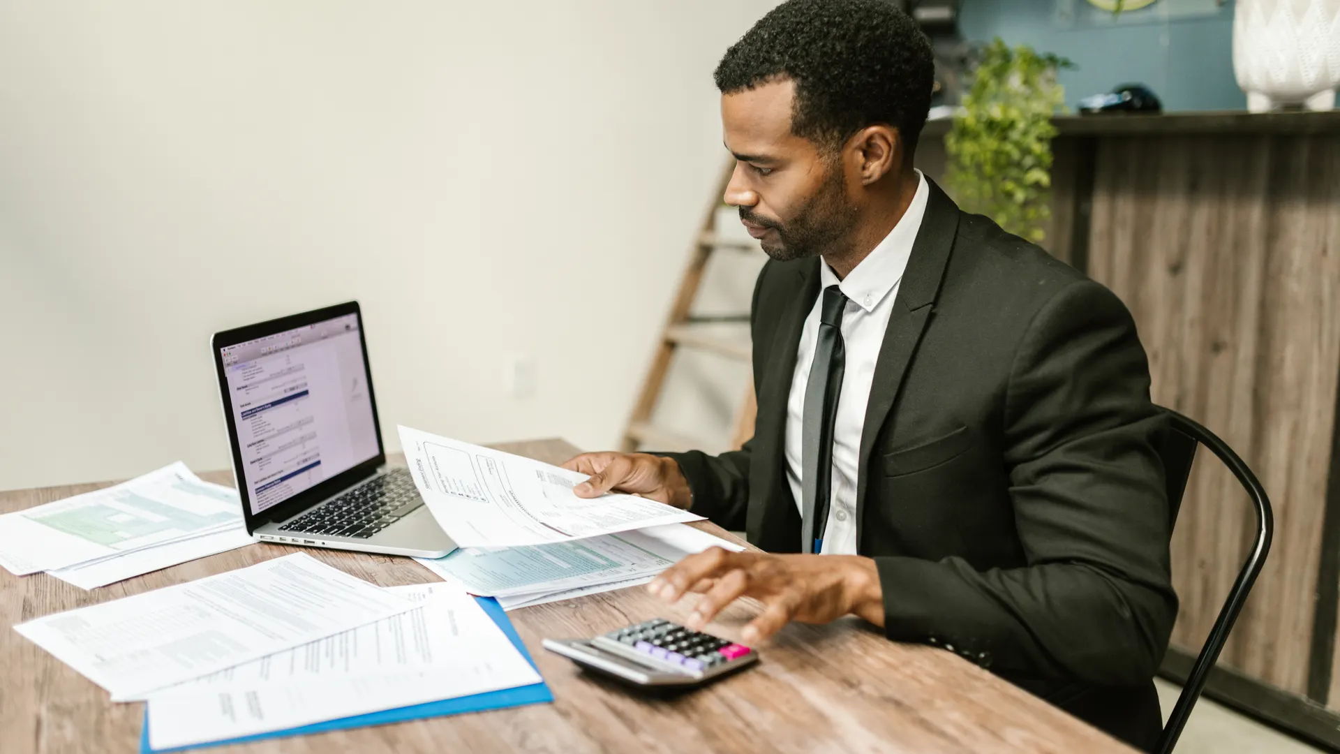 A businessman in a suit working on documents and using a laptop at an office desk