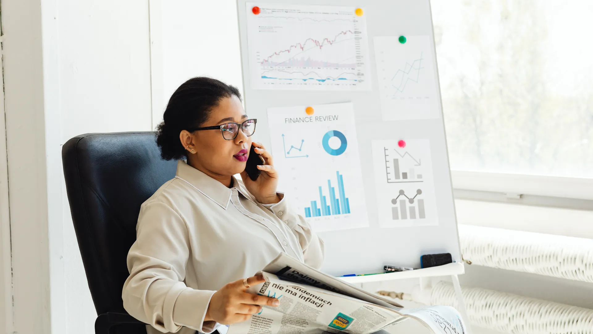 Businesswoman multitasking with phone and newspaper in office, examining financial charts