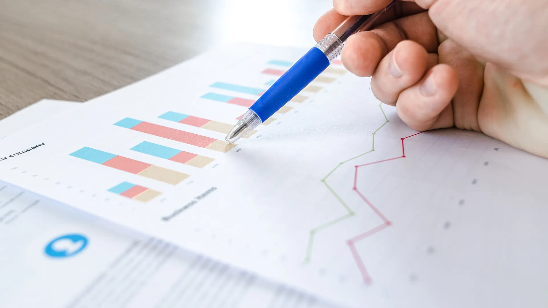 A close-up of a hand with a pen analyzing data on colorful bar and line charts on paper