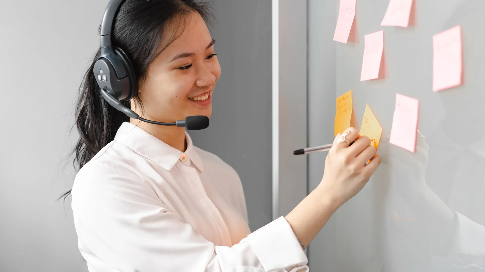 Young female call center agent wearing a headset, writing on sticky notes indoors