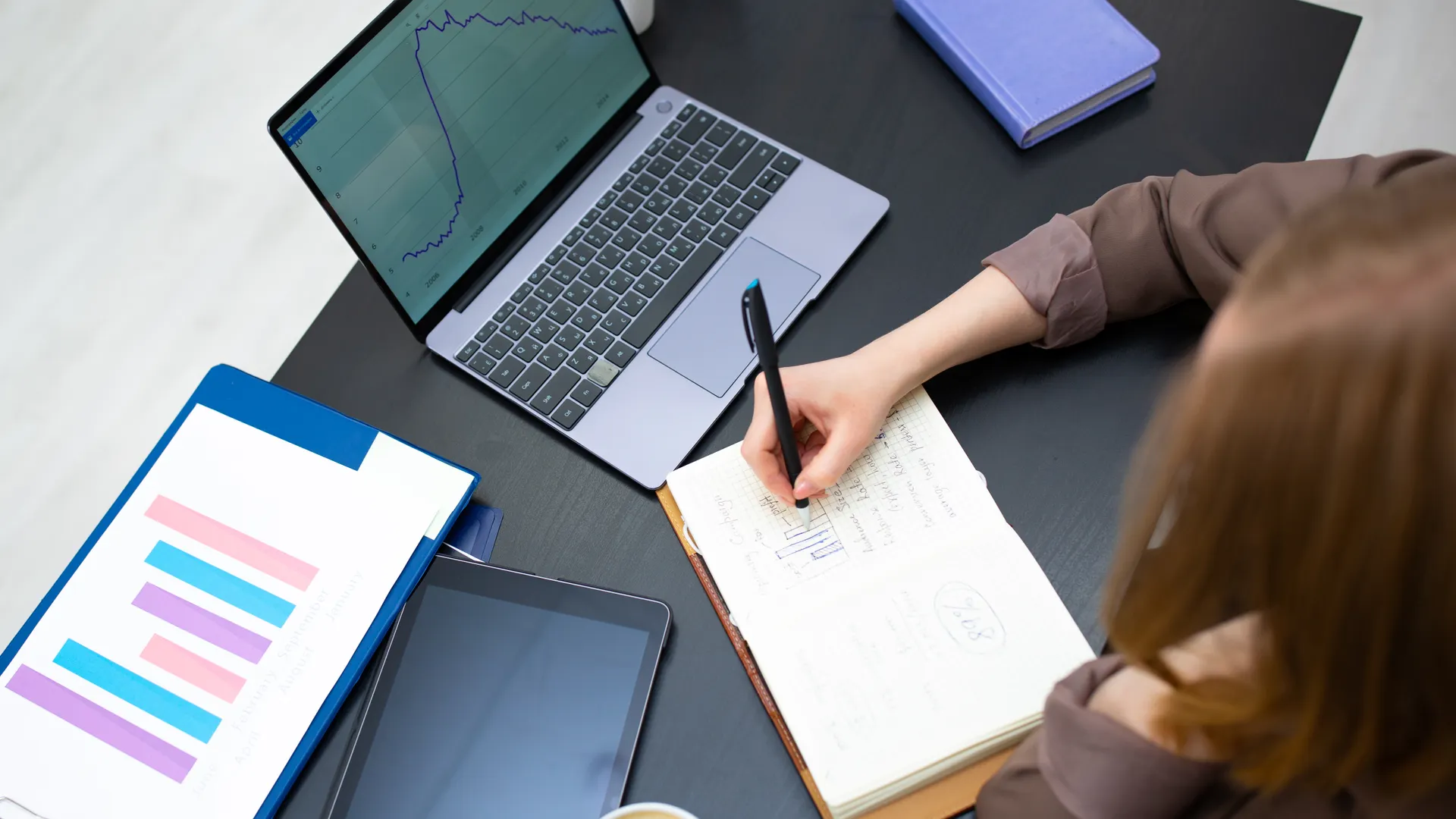 A person writing notes in a notebook beside a laptop displaying graphs in an office setting