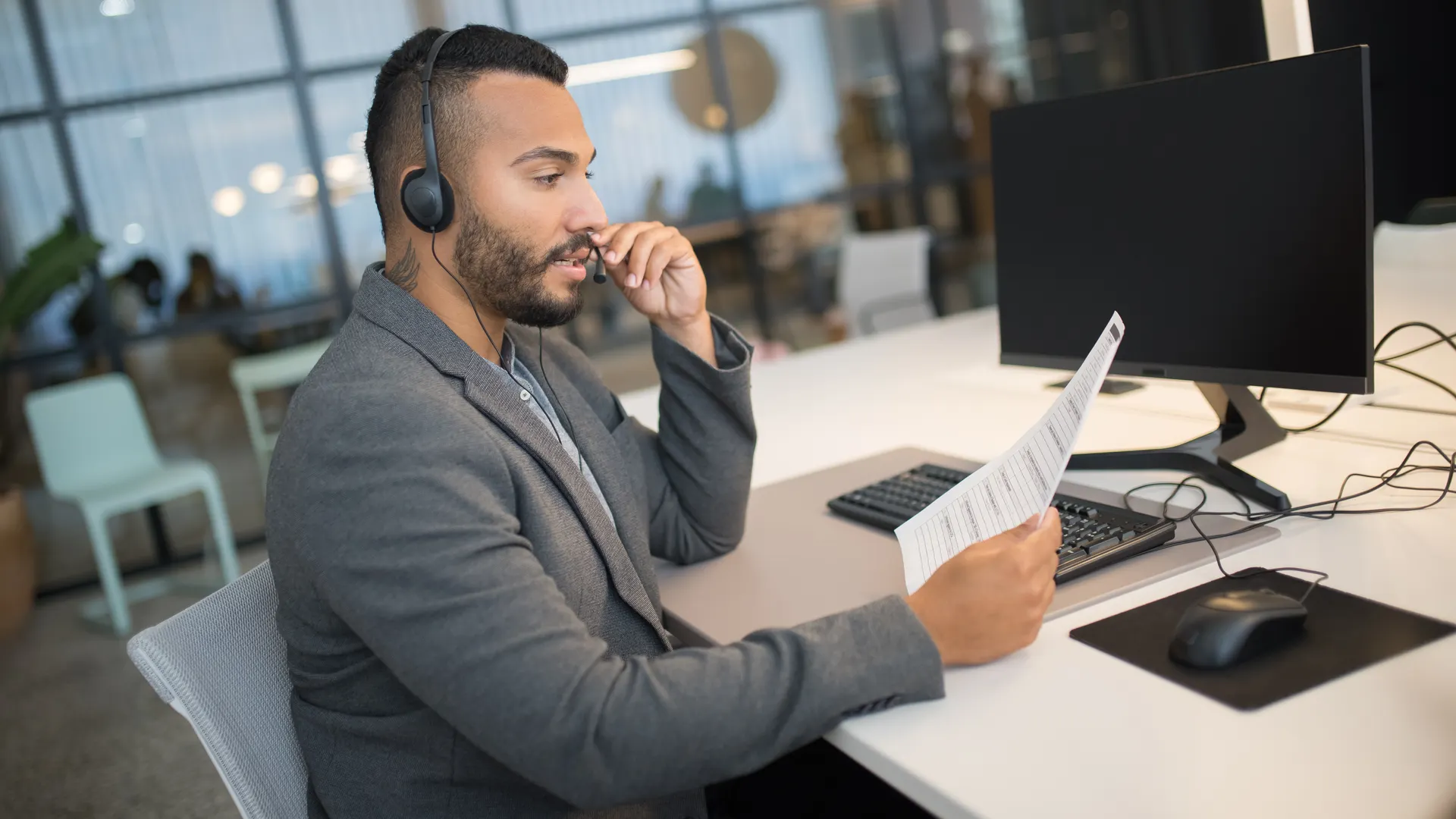 Young man in a call center, using a headset and reading documents at his office desk