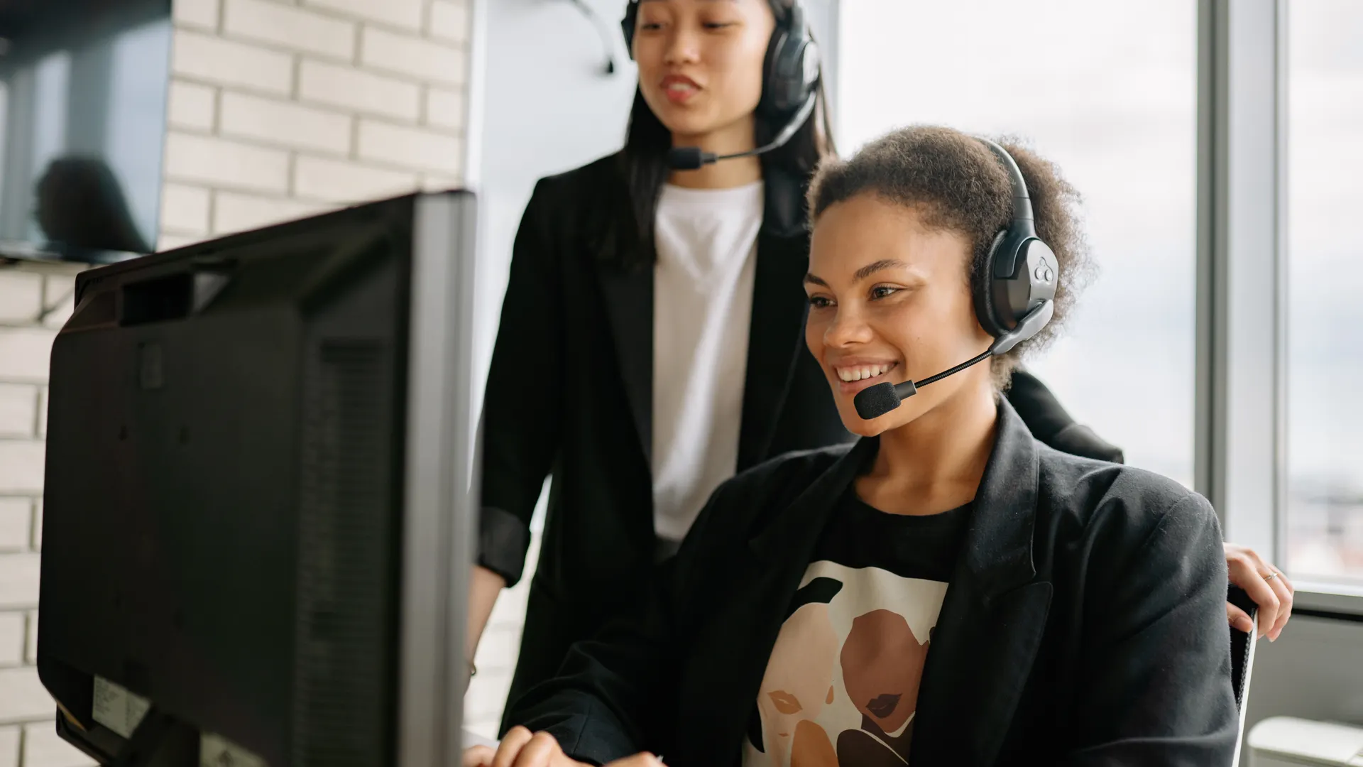 Two women with headsets engaged in customer service at a computer in a bright office
