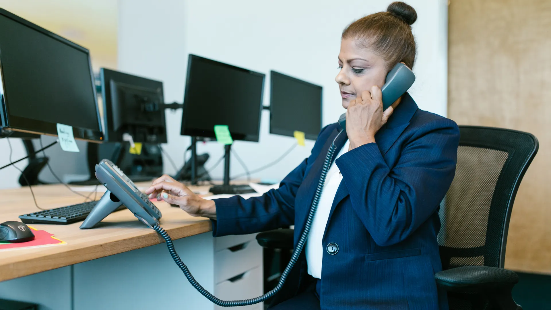 Businesswoman multitasking in office, using phone and computer, wearing formal attire