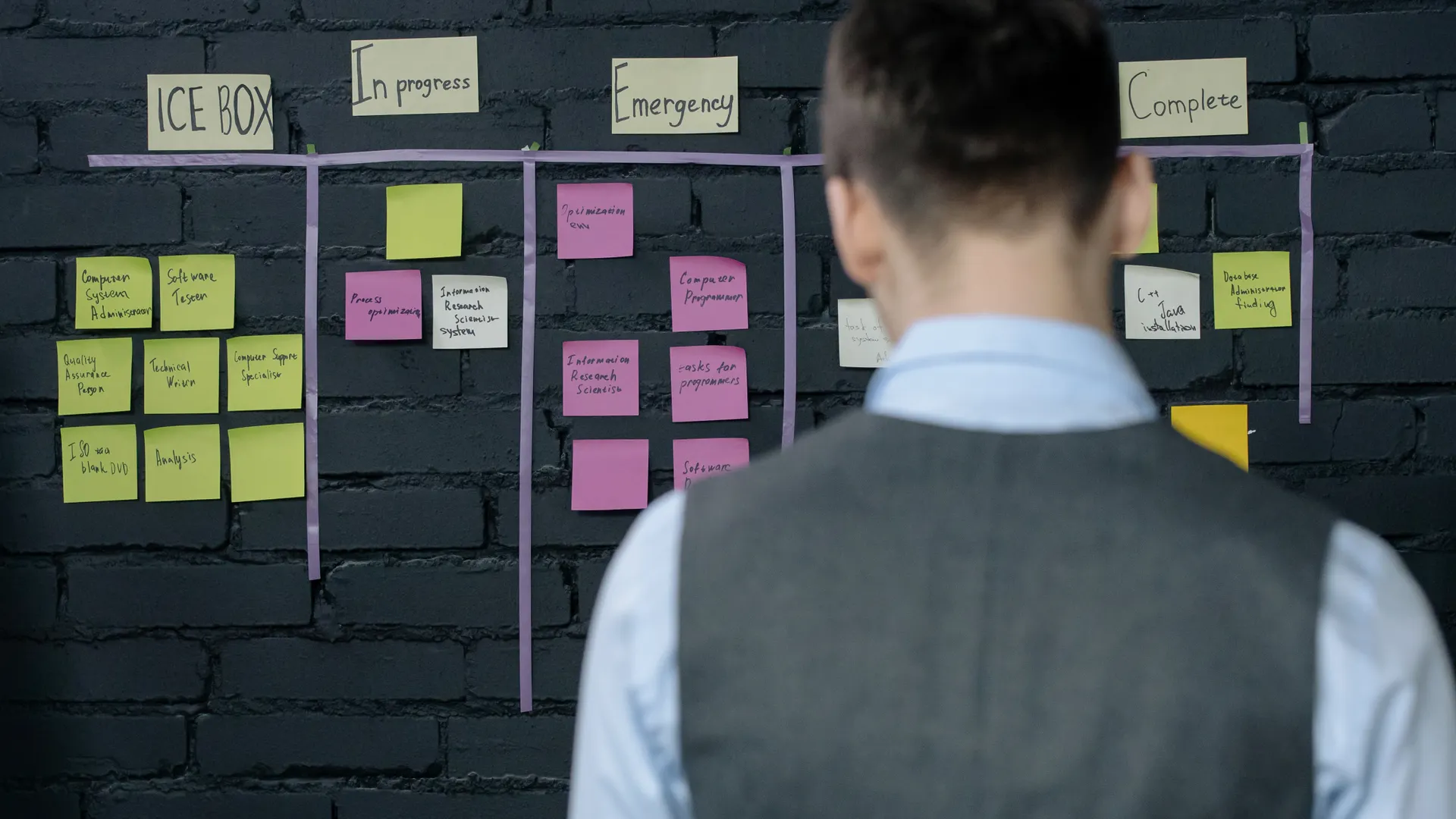 A person in a vest stands in front of a project management board with sticky notes on a dark brick