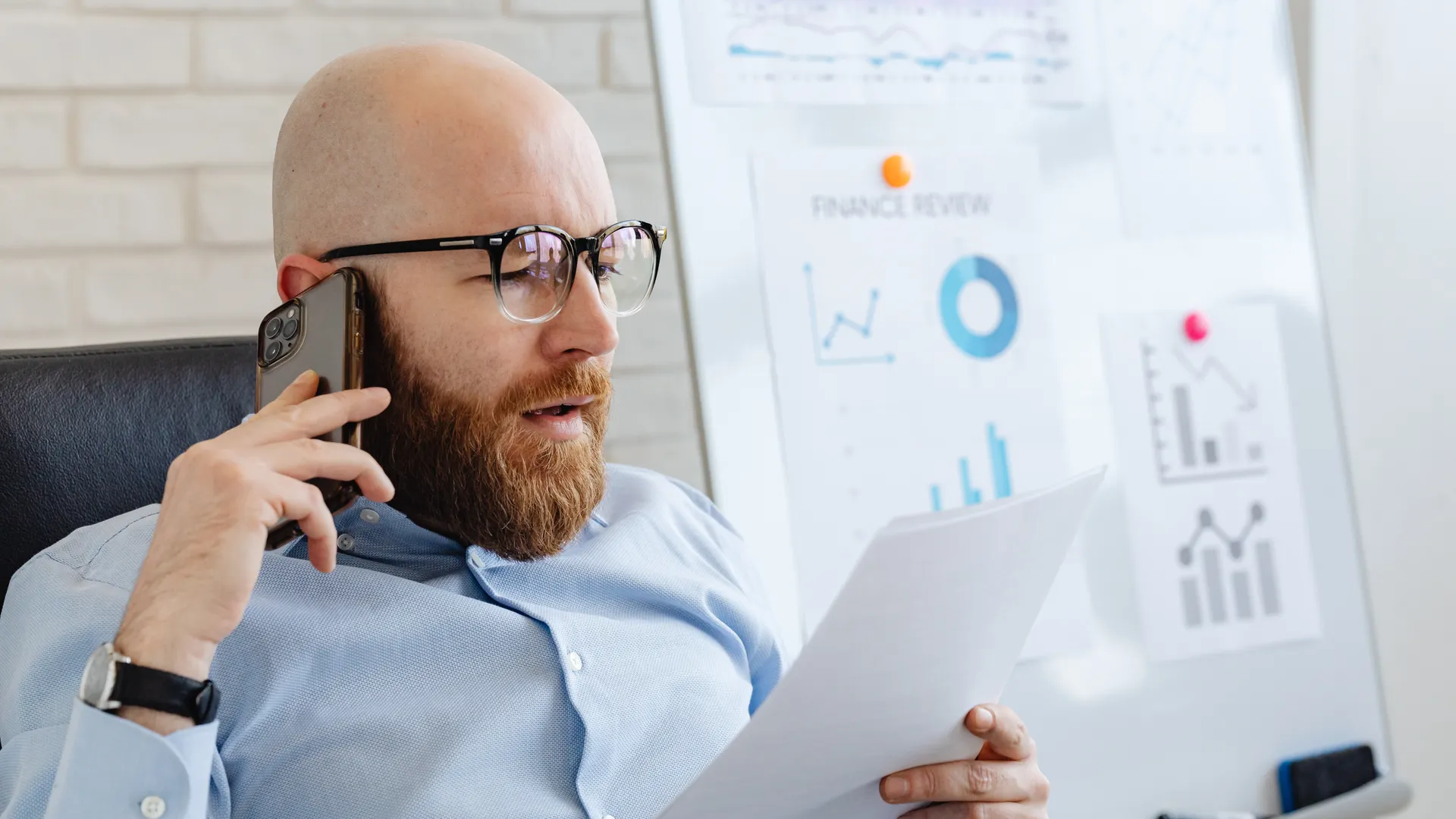 Bald man with beard holding smartphone and papers, reviewing financial charts in office