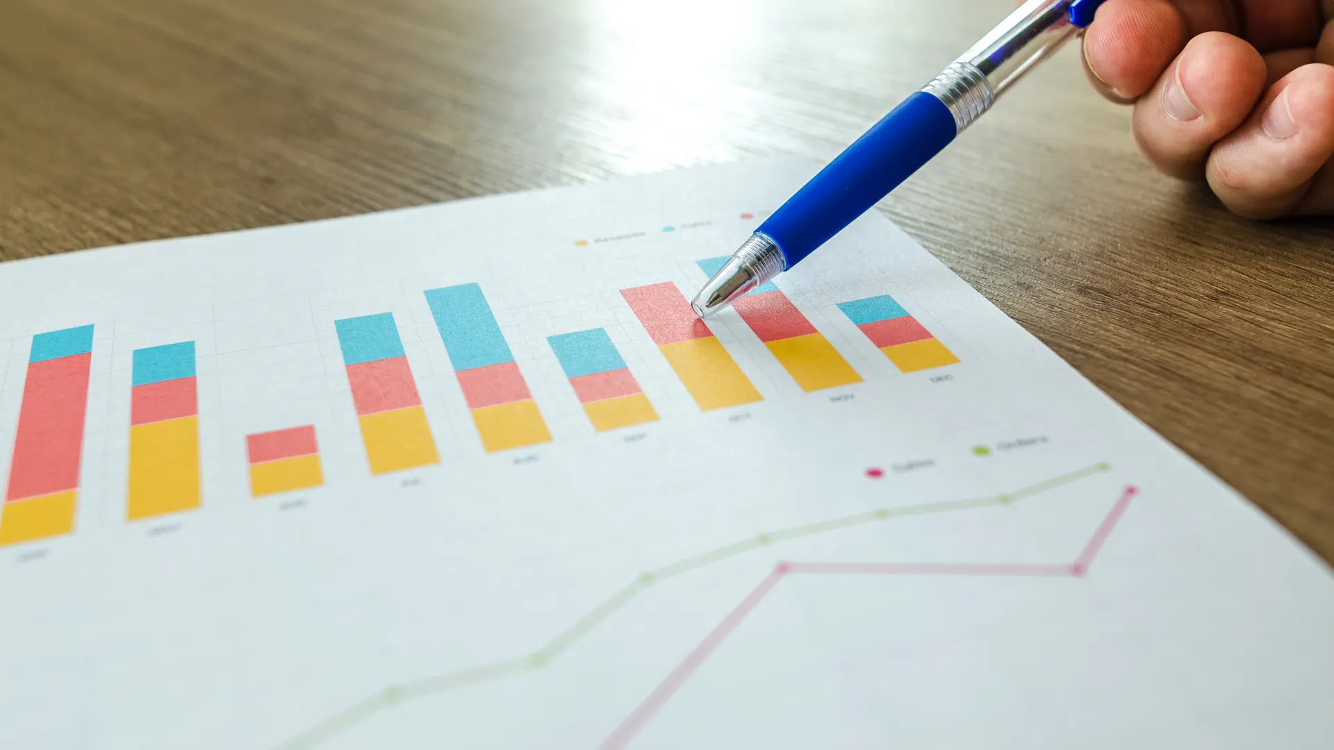 Detailed close-up of a hand pointing at colorful charts with a blue pen on wooden surface