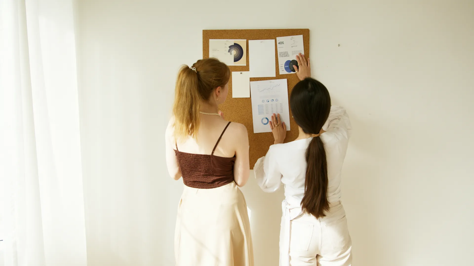 Two women analyzing charts and graphs on a corkboard. Business teamwork concept