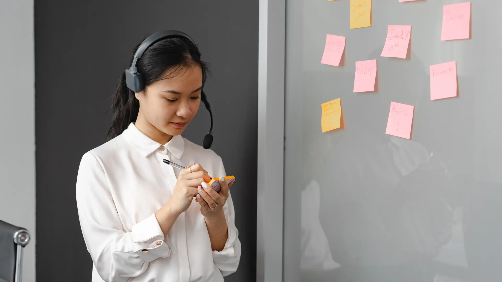 A woman in a call center writing on sticky notes with a headset indoors
