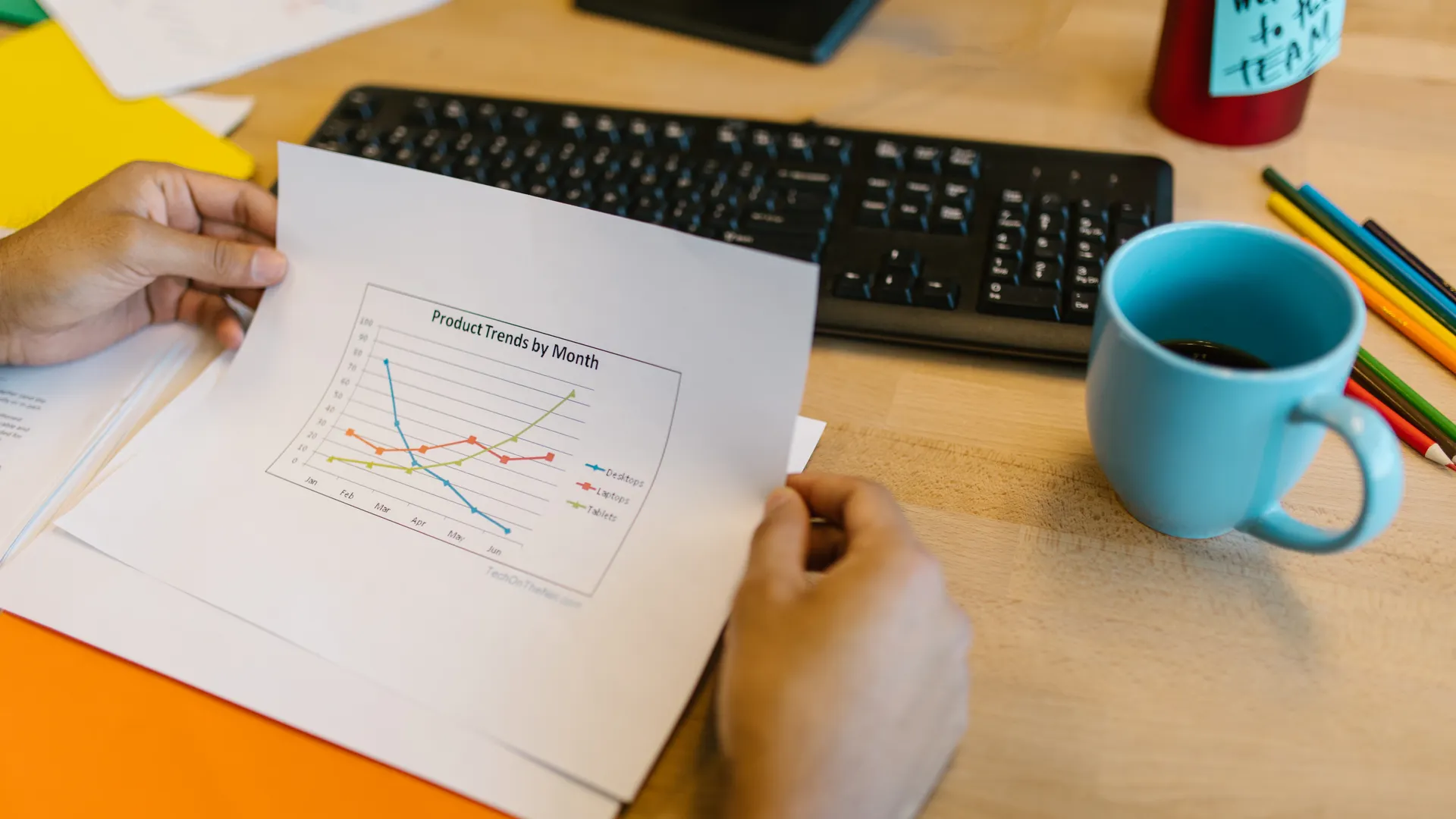 Close-up of hands reviewing a paper graph with a coffee cup and keyboard on a desk
