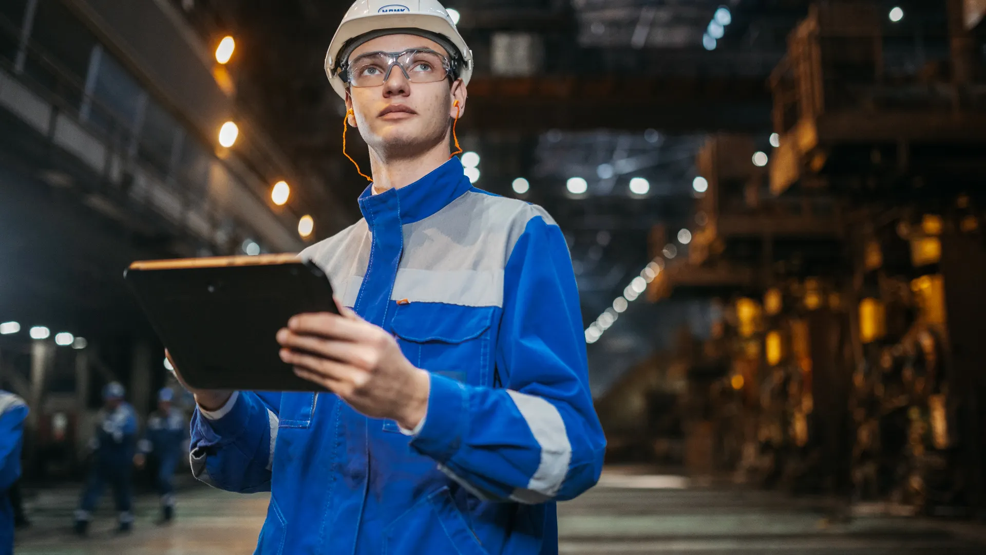 Engineer in safety gear uses a tablet in a large industrial factory setting, focused and diligent