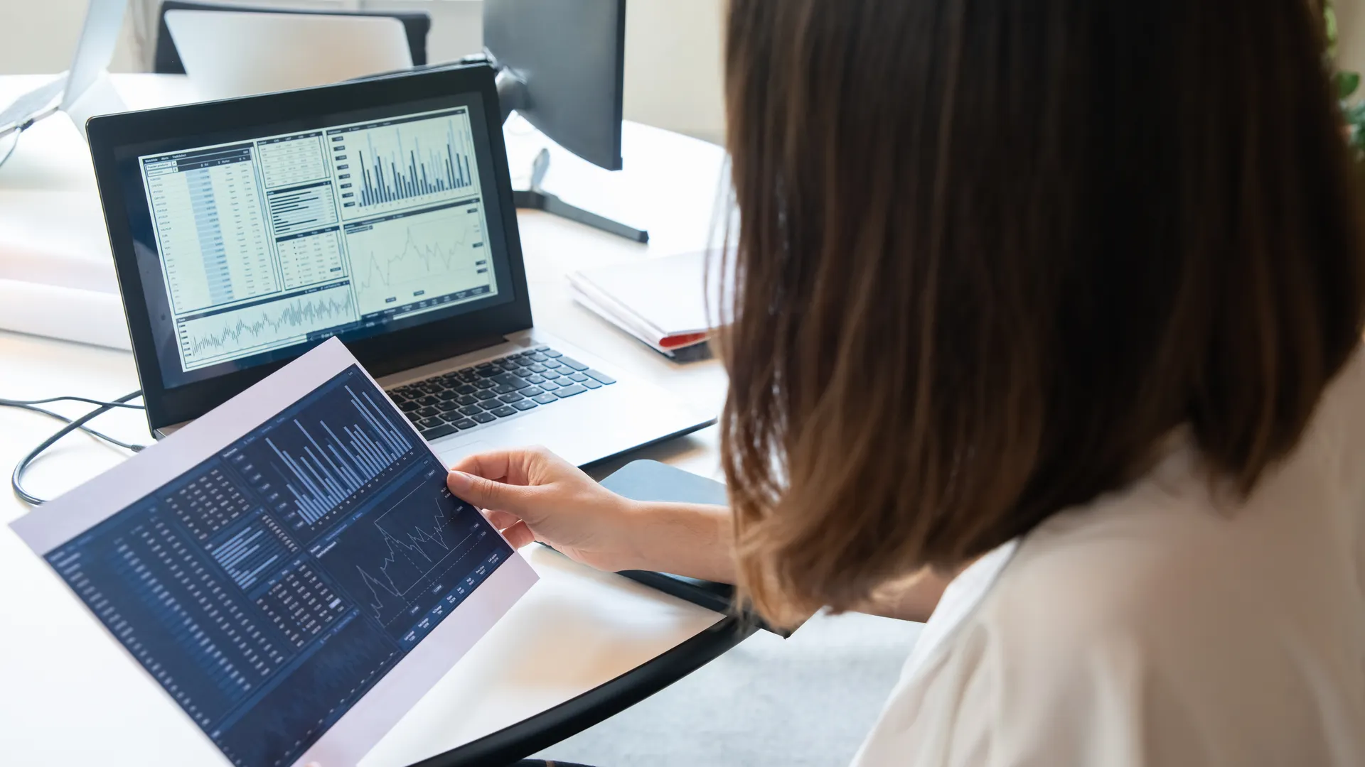 A professional woman analyzes financial charts on a laptop and paper for business insights