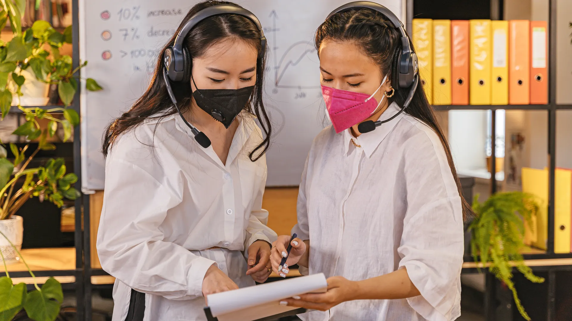 Two women in a modern office reviewing documents while wearing masks and headsets