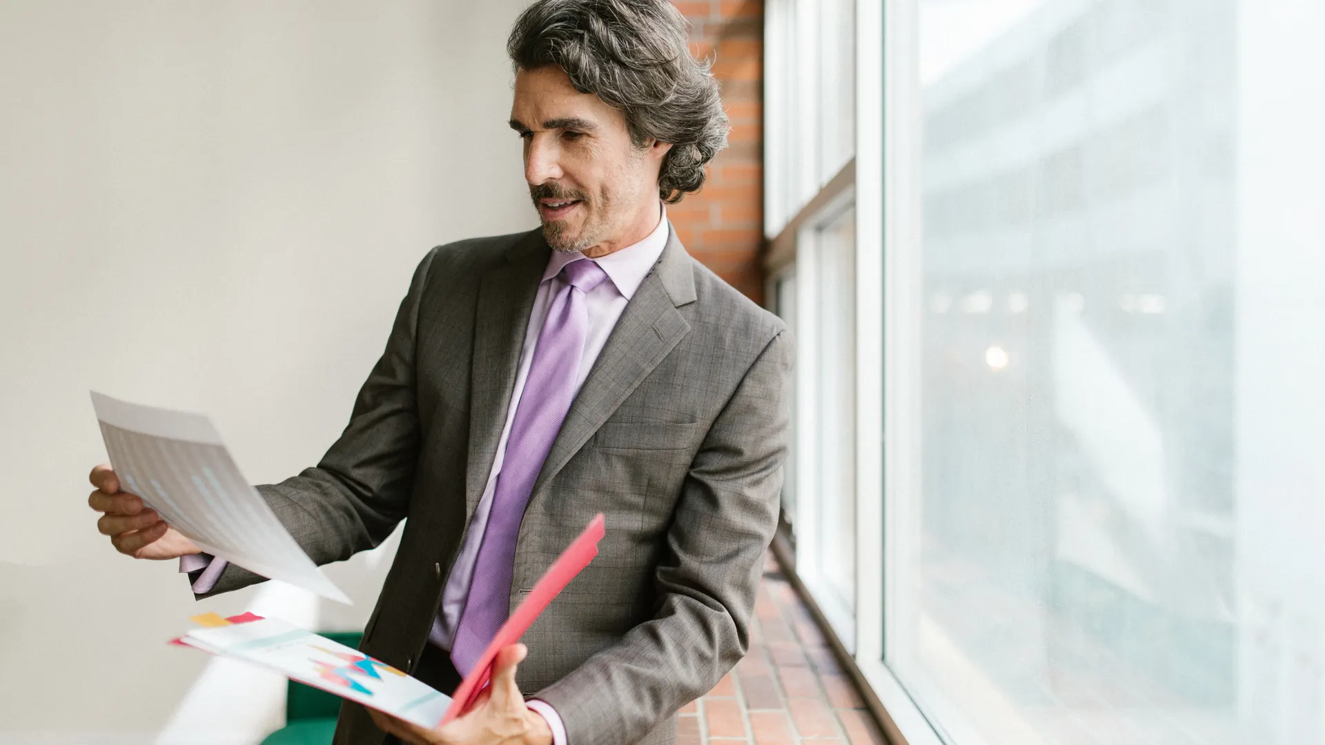 Businessman in a gray suit reviewing paperwork by a large window in an office setting