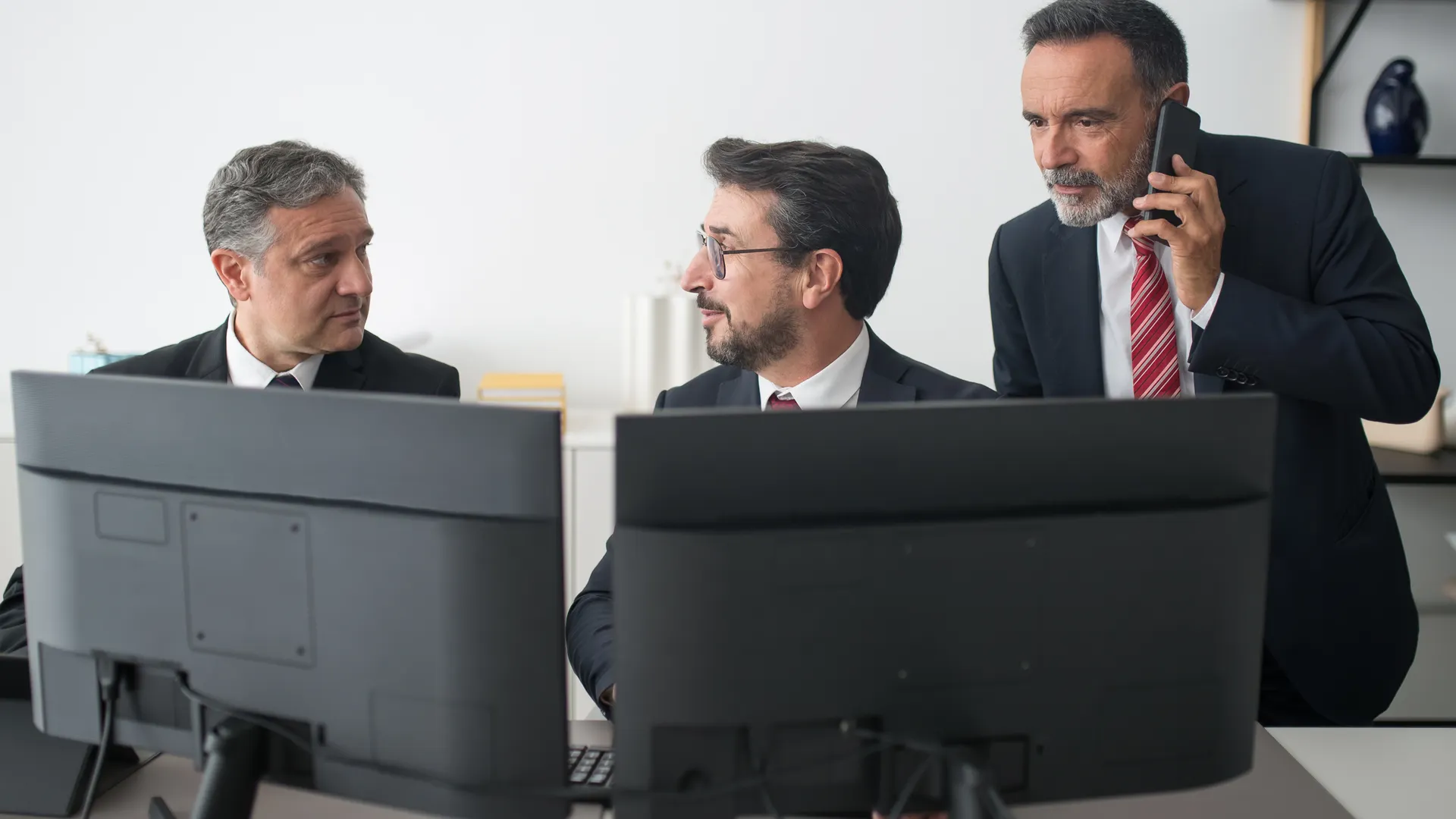 Three businessmen in an office discussing work, two using computers and one on a phone call