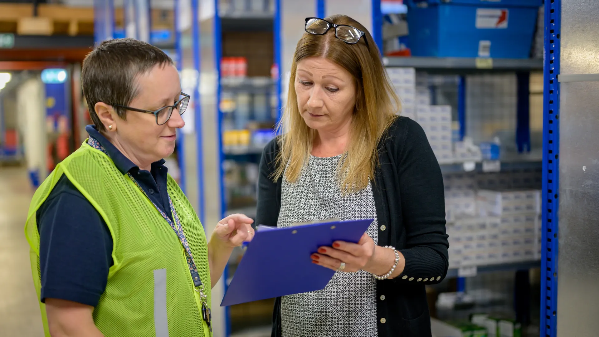 Two women in a warehouse reviewing an inventory checklist on a clipboard