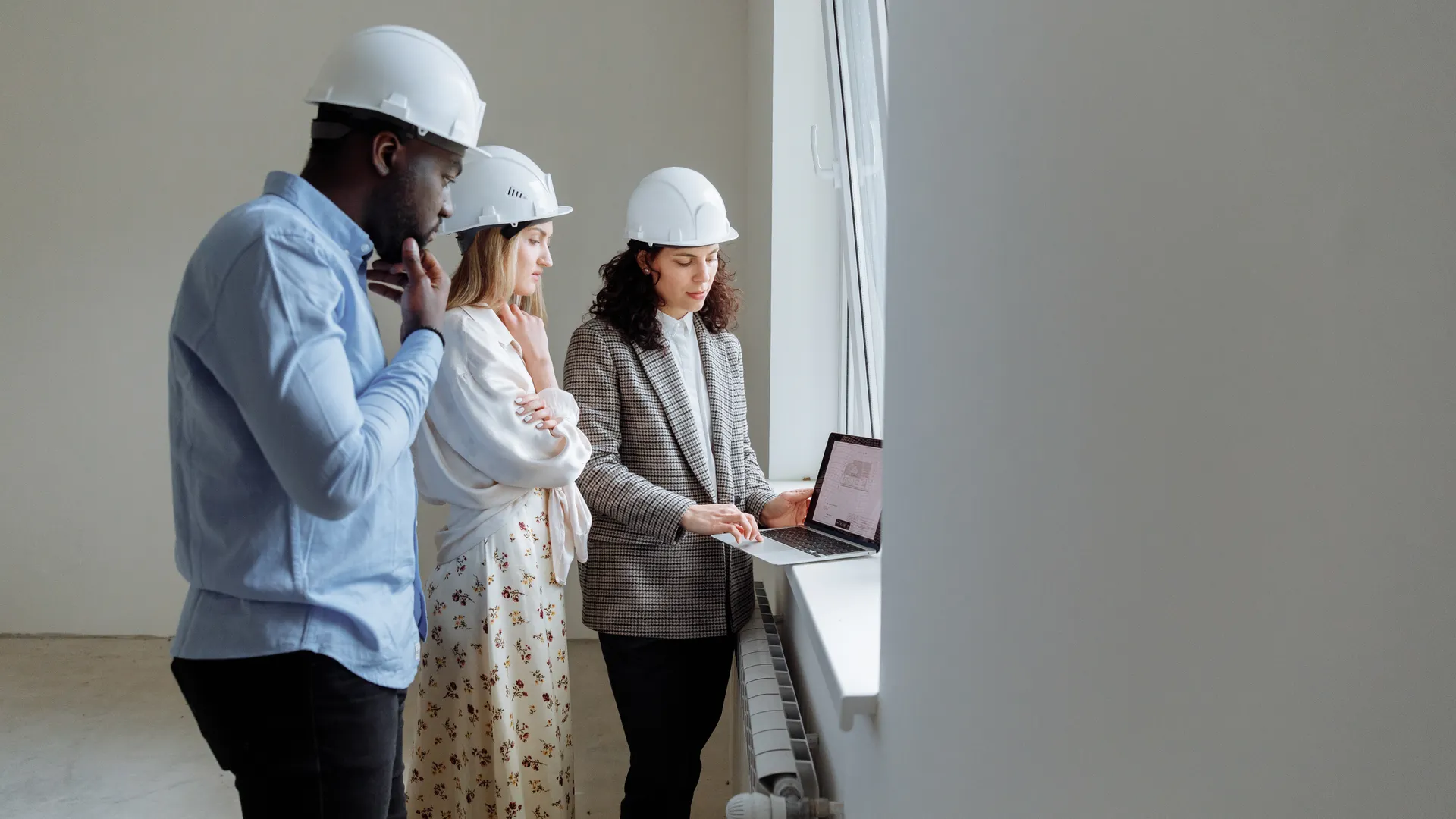 Three engineers wearing helmets review building plans on a laptop in a bright indoor setting