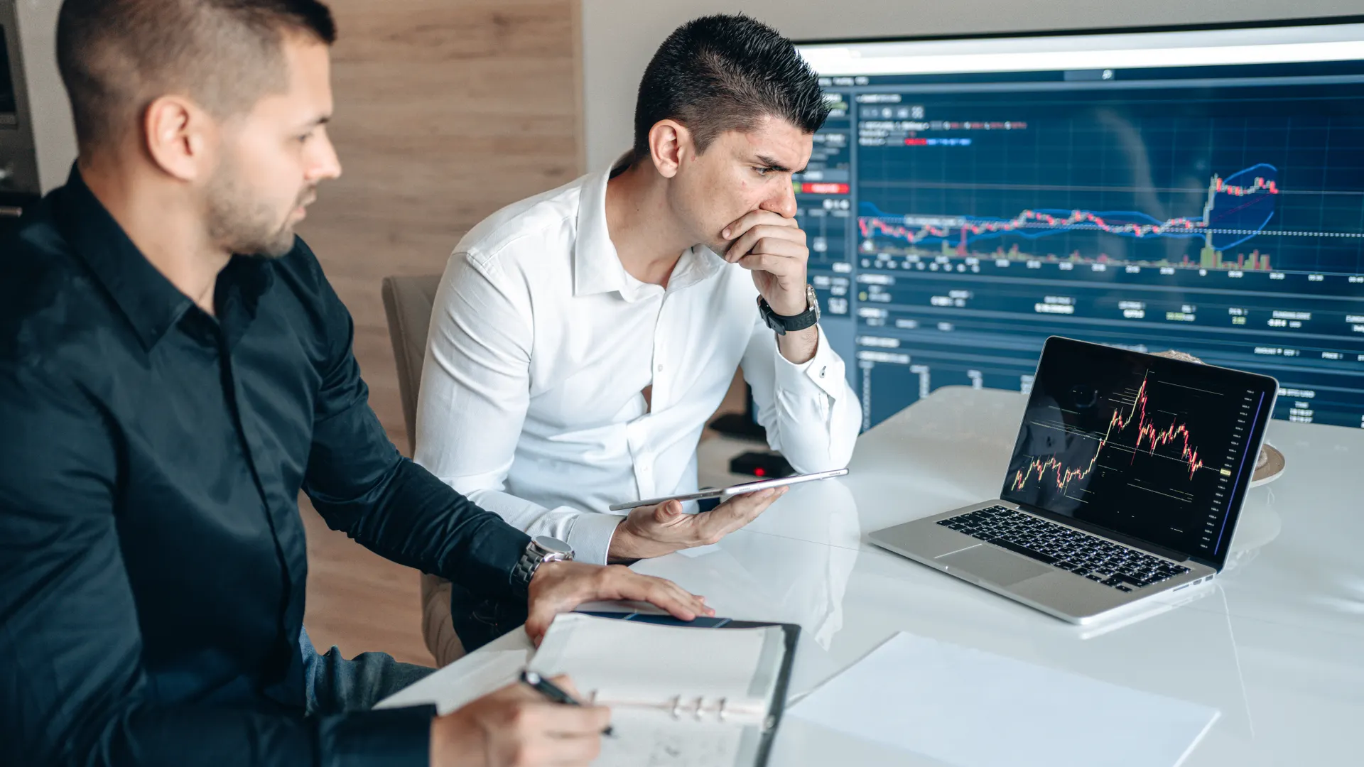 Two businessmen analyzing stock market data on laptops and tablets in an office setting