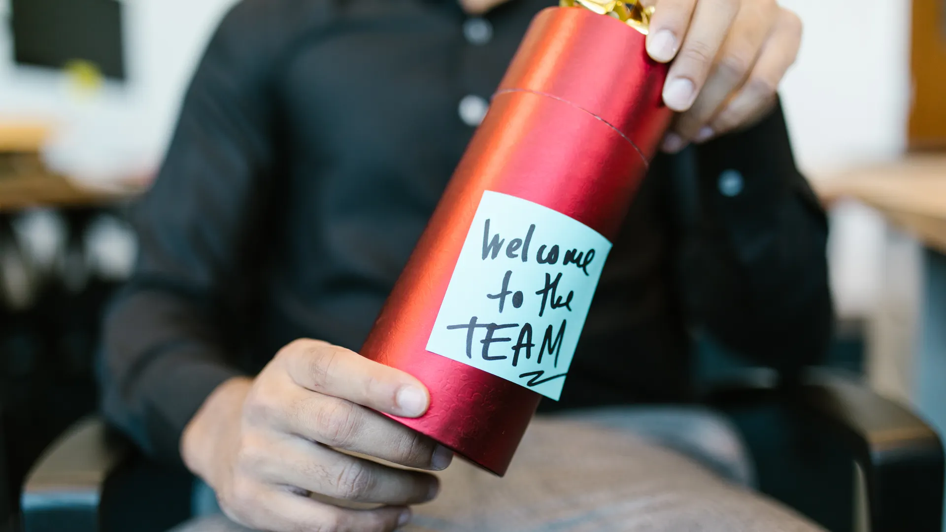 A red gift box held by a person with a welcome to the team note, ideal for new hire announcements